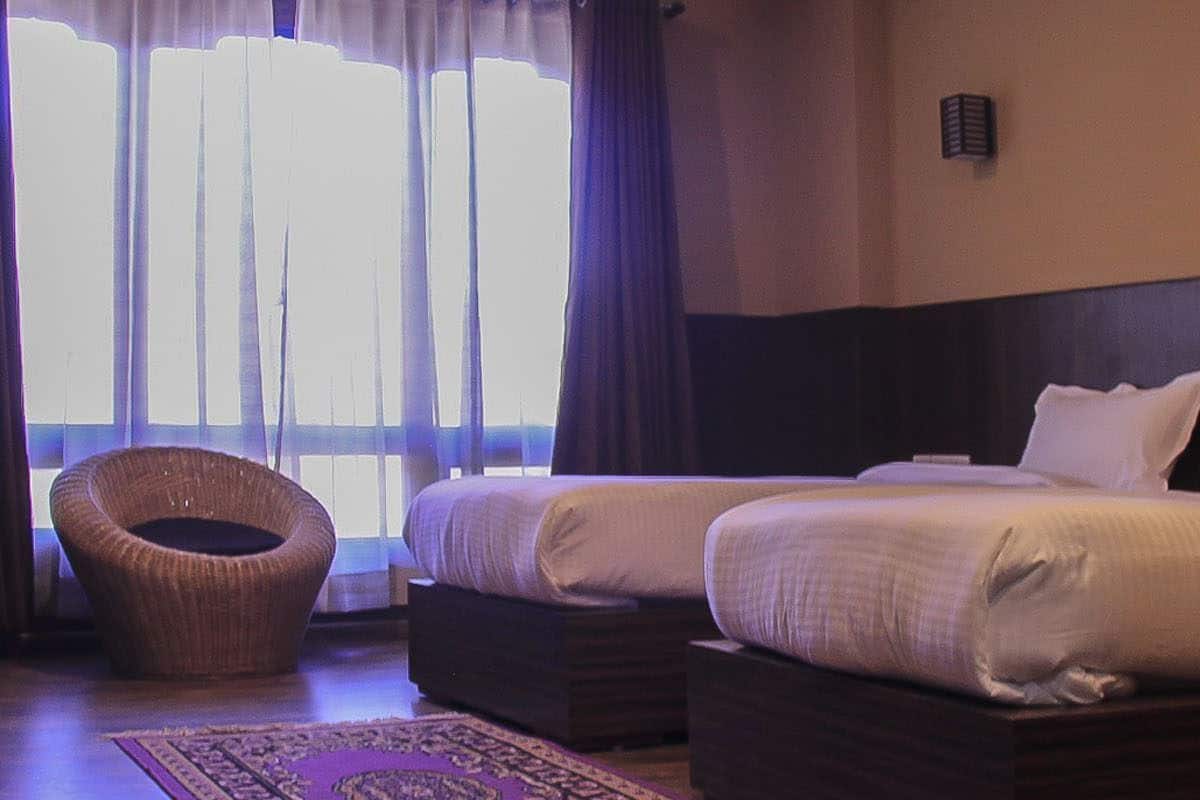 Une chambre d’hôtel avec deux lits, un fauteuil en osier et une grande fenêtre avec des rideaux.