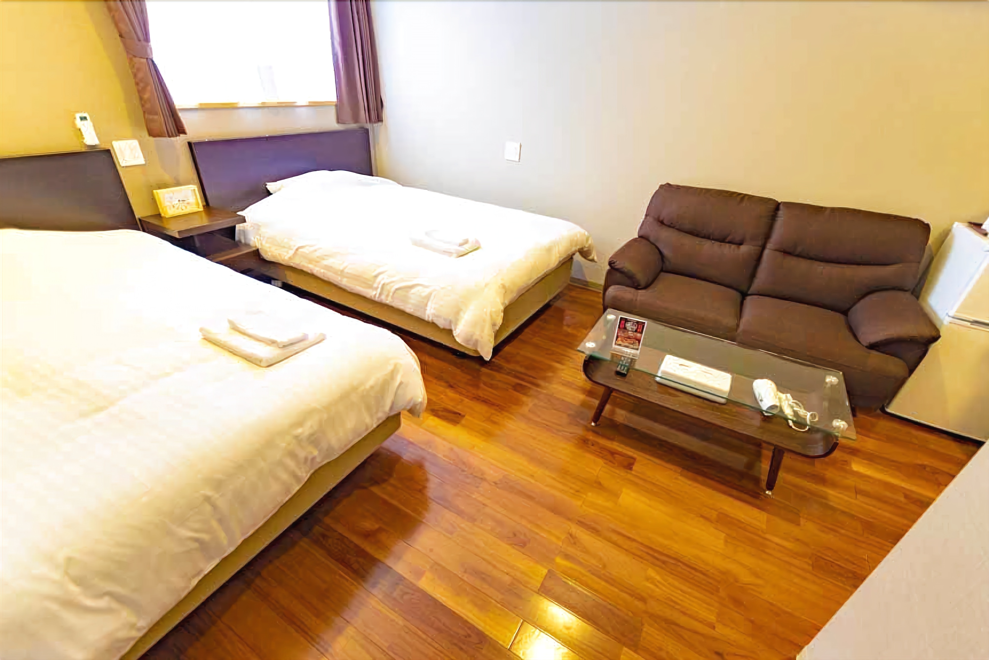 Habitación de hotel con dos camas individuales, un sofá marrón, una mesita con un control remoto y una ventana con cortinas.