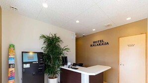 Un hall d'hôtel comprenant un comptoir d'accueil, une plante, un distributeur automatique et une enseigne murale indiquant « HÔTEL KALAKAUA ».