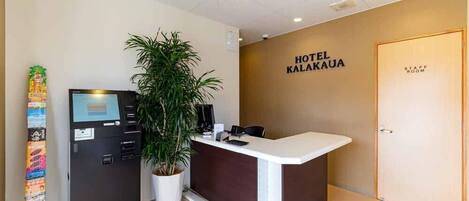 A hotel reception area with a reception desk, a plant, a vending machine, and a wall sign that reads "HOTEL KALAKAUA".