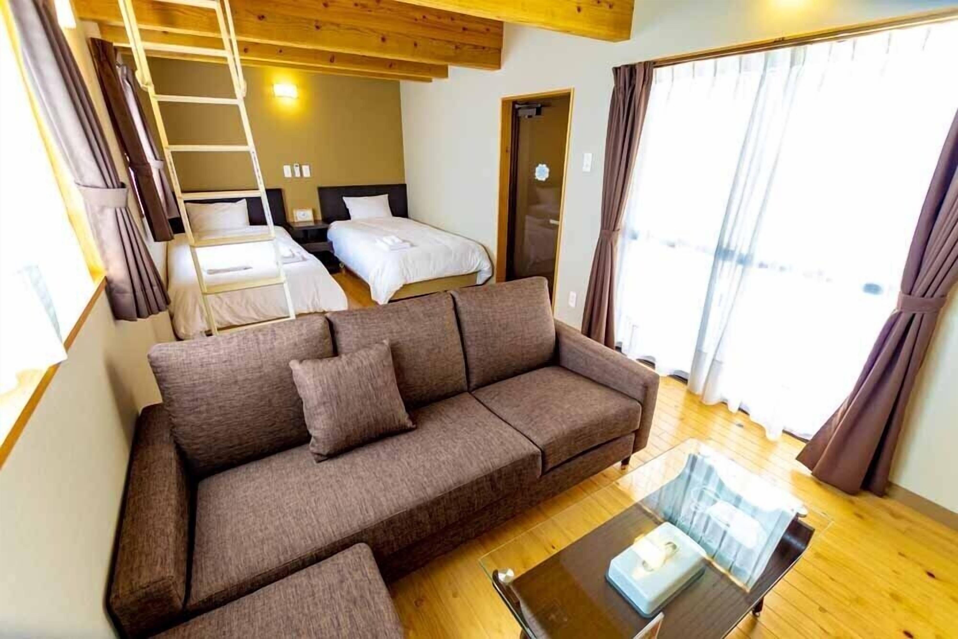 A room with a brown sofa, a glass coffee table, a bed with white bedding, and a wooden ceiling.