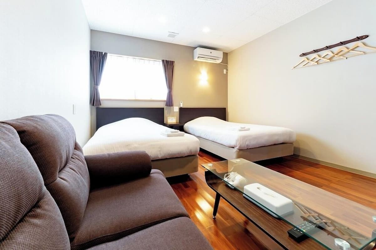 Habitación de hotel con sofá, dos camas, una mesa de centro de vidrio, un equipo de aire acondicionado y un perchero en la pared.