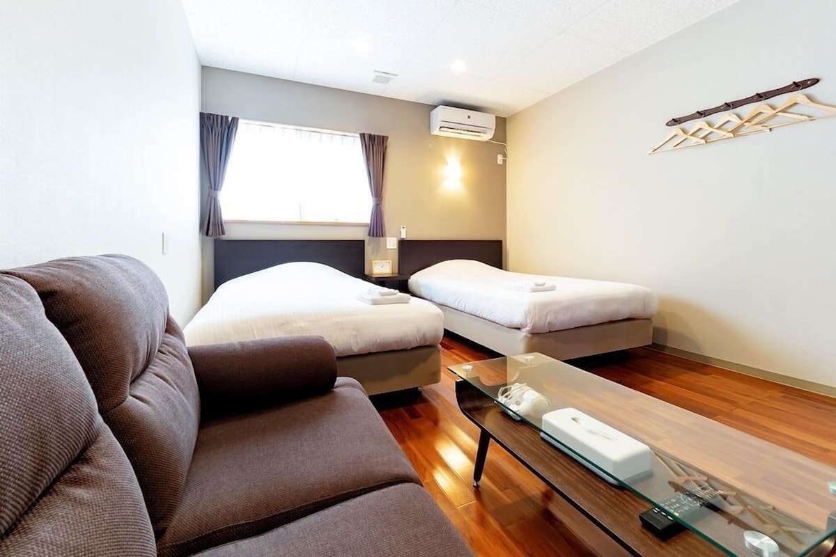 A modern hotel room with a sofa, two single beds, a glass coffee table, and an air conditioning unit.