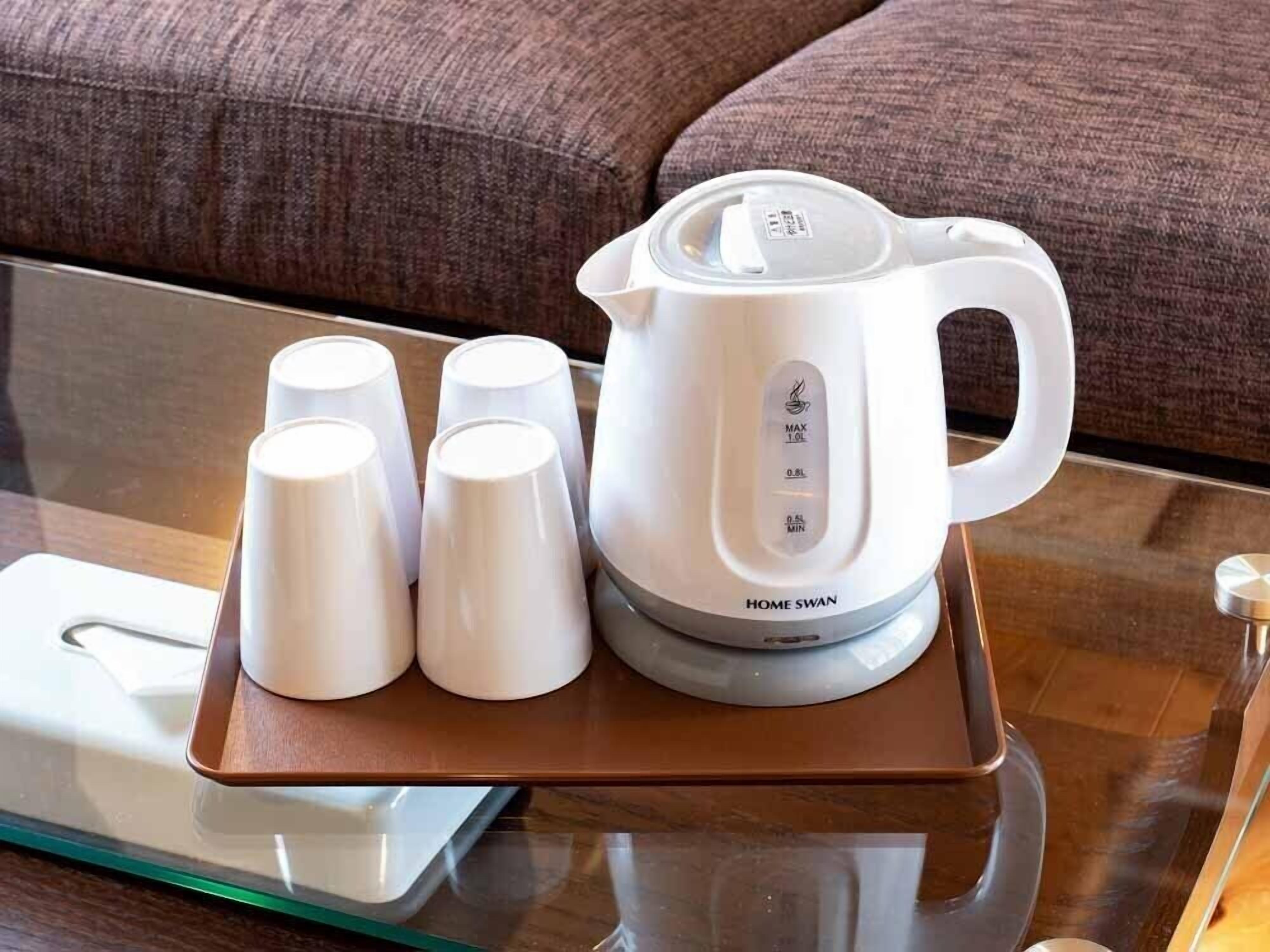 A white electric kettle with a tray, four white cups, and a remote control on a glass table.