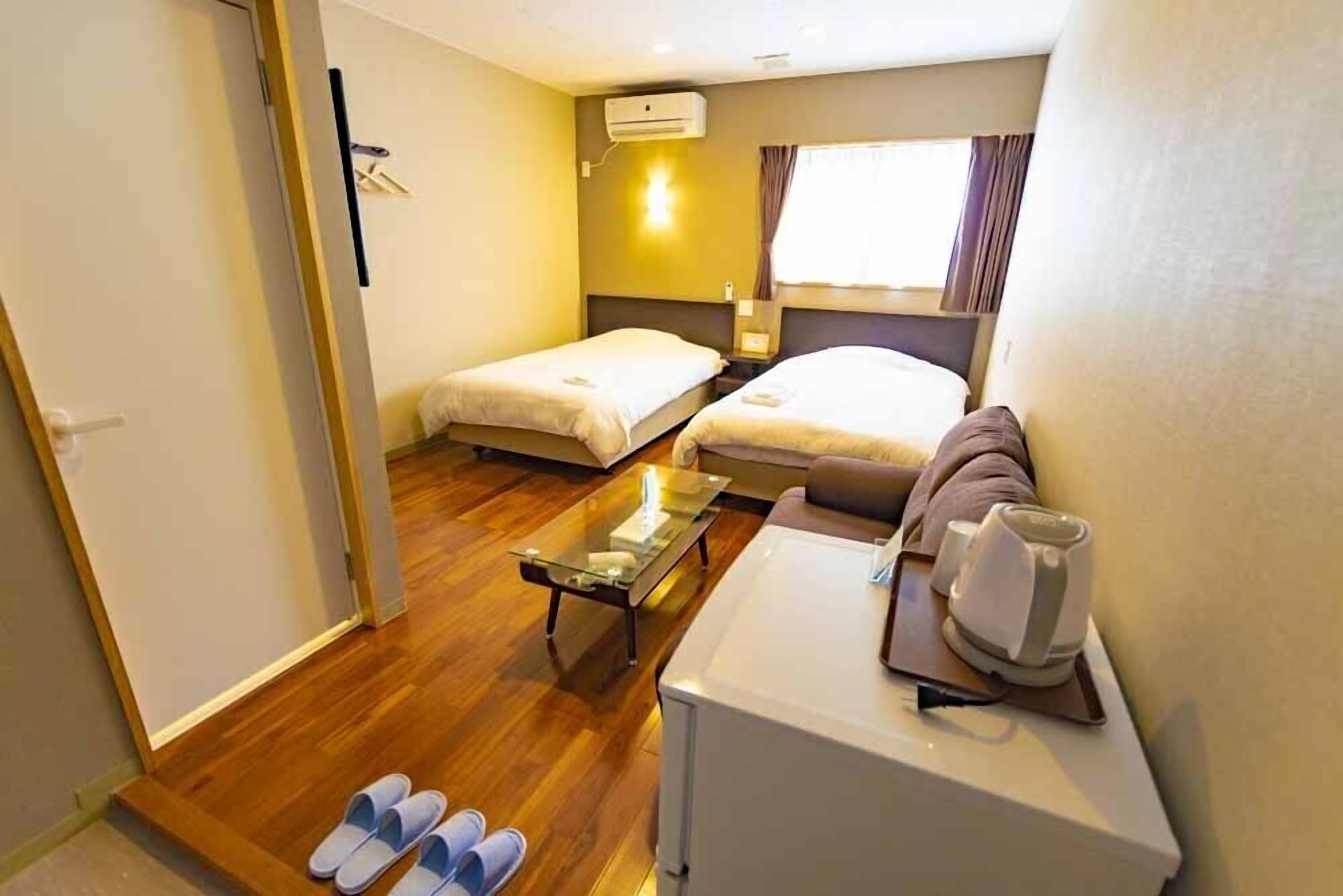 A hotel room with two beds, a sofa, a coffee machine, and slippers on the floor.