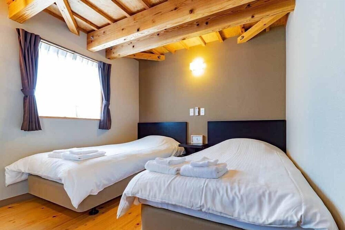 A room with two single beds, wooden flooring, a sloped ceiling with exposed beams, a window with curtains, and a wall-mounted light.