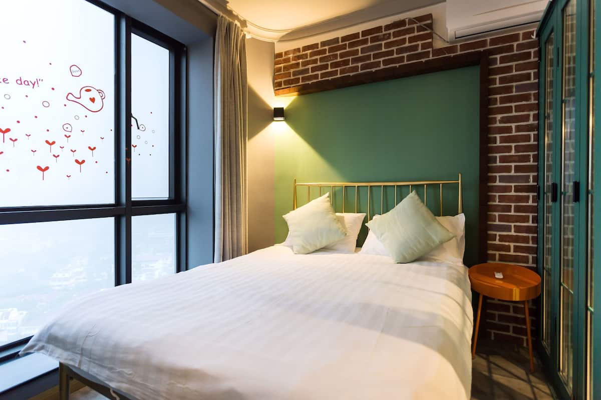 Ein Hotelzimmer mit großem Fenster, einem Bett mit Kissen, einem runden Holfnstisch und einer Backsteinwand.