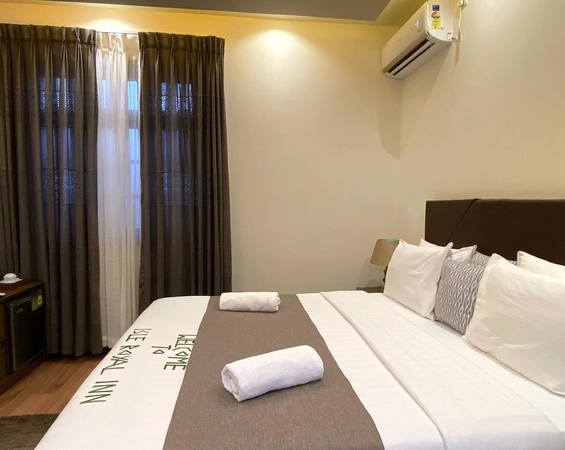A hotel room with a bed, a nightstand, a lamp, an air conditioning unit, and a window with curtains.