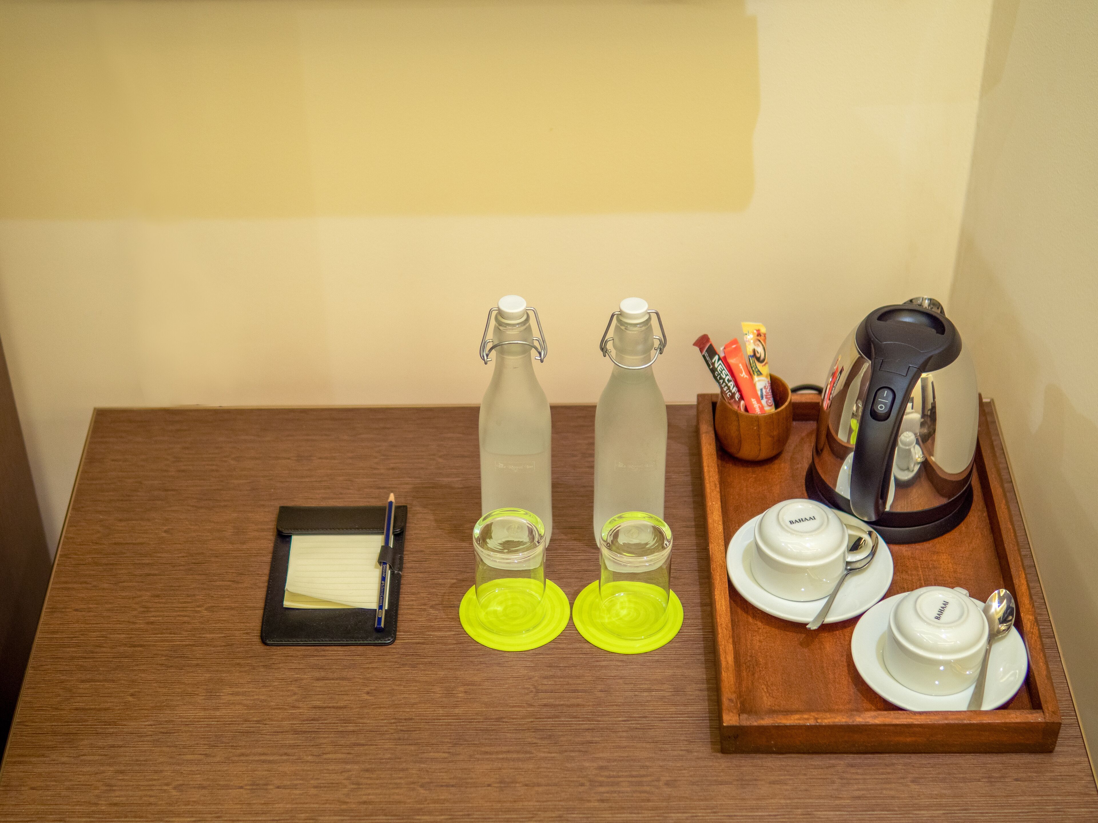 Una mesa de madera con un cuaderno, dos botellas de agua, un vaso, una cafetera y una bandeja con tazas y una cuchara.