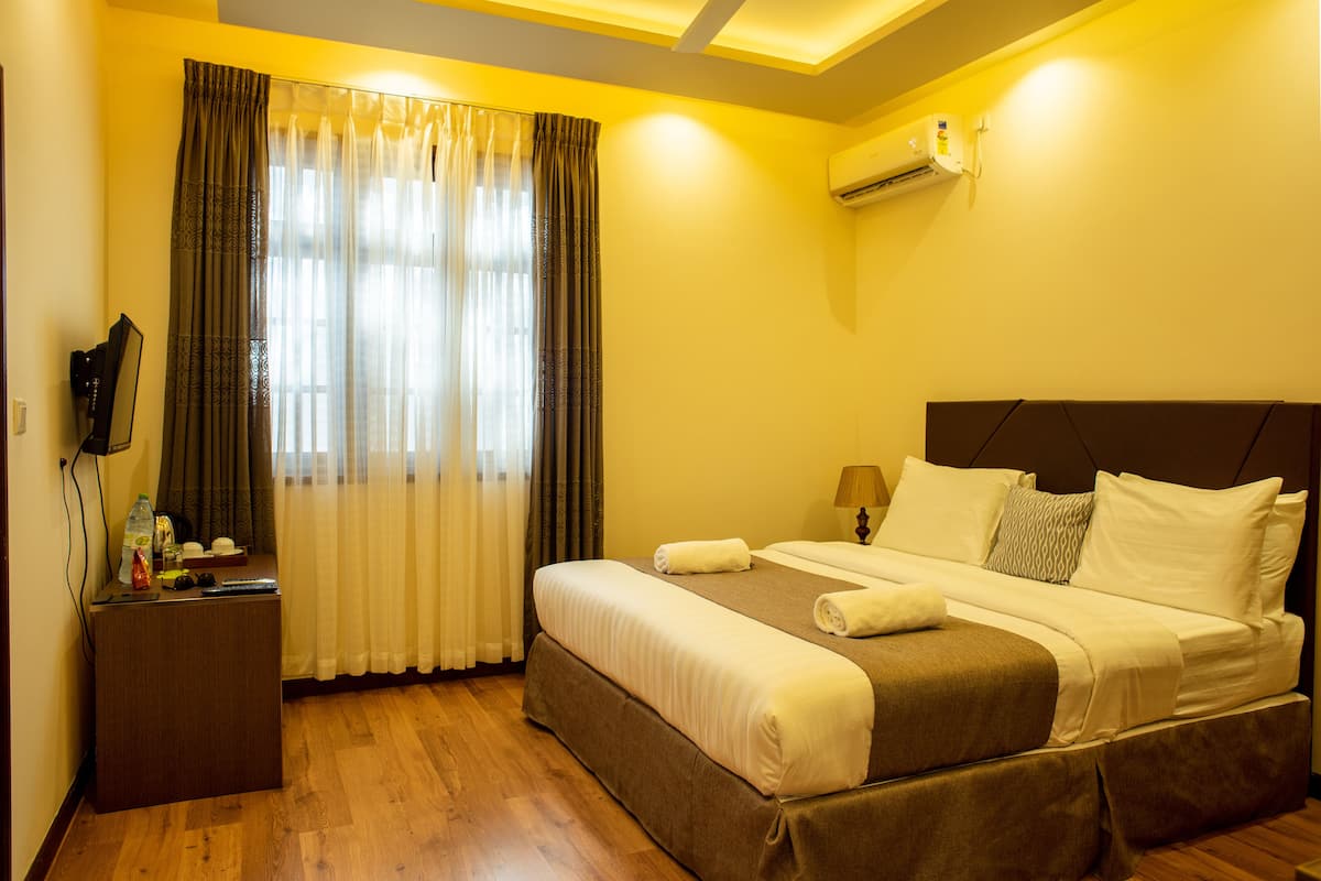 A hotel room with a bed, a bedside table, a lamp, curtains, and an air conditioning unit.