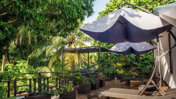 A wooden deck with a lounge chair and umbrella, surrounded by lush greenery and potted plants.