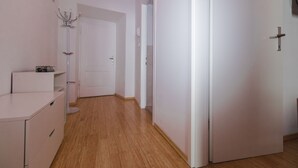 Un pasillo con suelo de madera, paredes blancas y una puerta de color blanco. Se ve un perchero y una pequeña mesa con un marco de fotos a la izquierda.