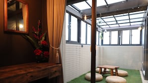 A cozy room with a wooden bench, a mirror, and a vase with red flowers. There is a window with a view of the sky and a hanging light fixture. - Song Qian Brigade (Lukang)
