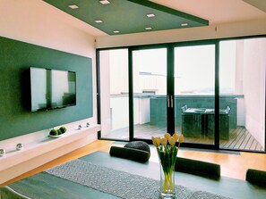 Uma sala de estar moderna com televisão de tela plana, uma mesa de centro de vidro e vista para o exterior por meio de grandes portas deslizantes.