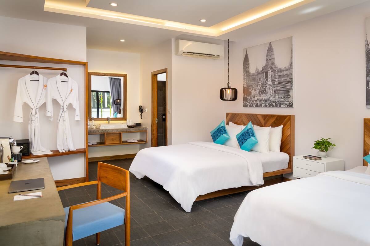 A hotel room with two beds, a desk with a laptop, a chair, a mirror, a vanity with a sink, and artwork on the walls.