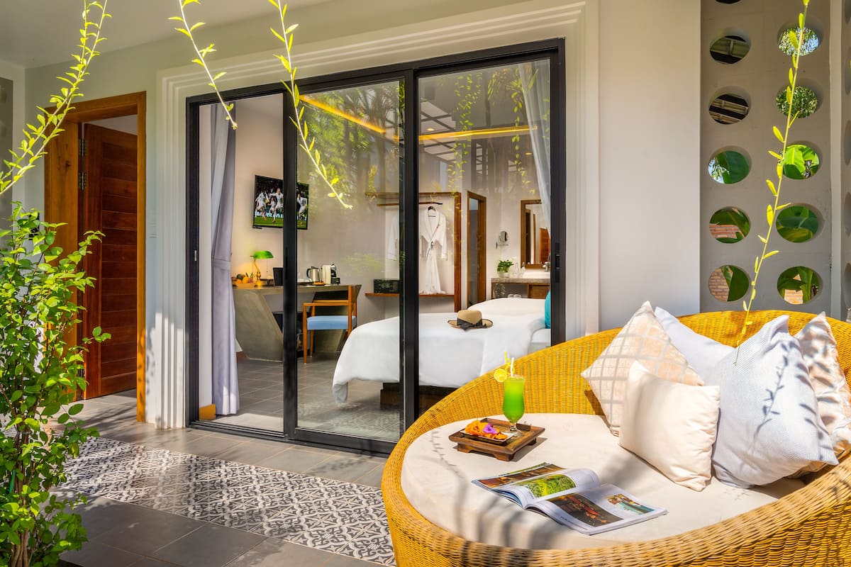 A modern hotel room with a large bed, a wicker sofa with a drink, and a view through a glass door.