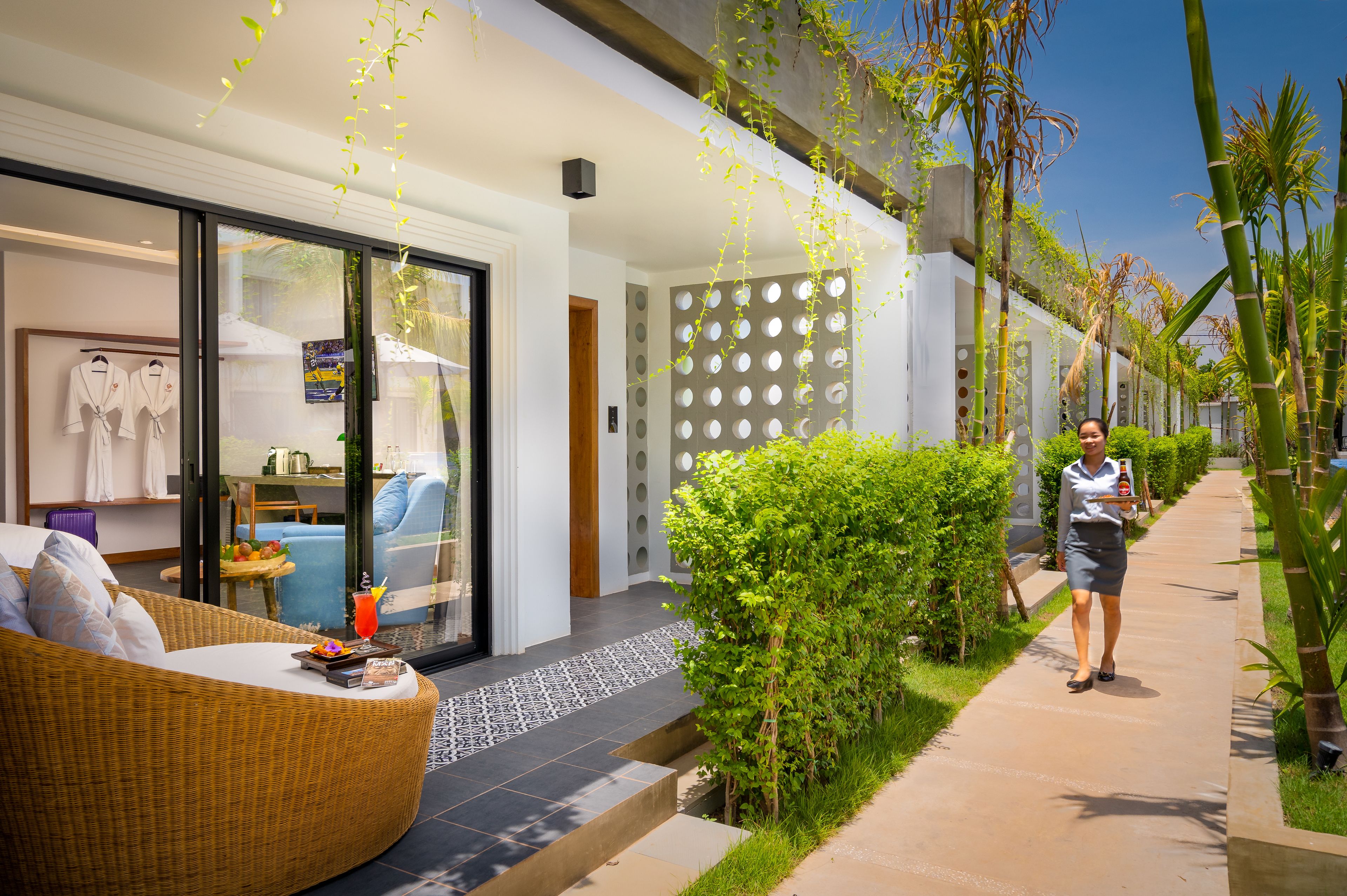 L’extérieur d’un hôtel moderne, avec une entrée dotée d’une porte en verre, un espace détente aménagé avec des meubles en osier et un chemin bordé de verdure.