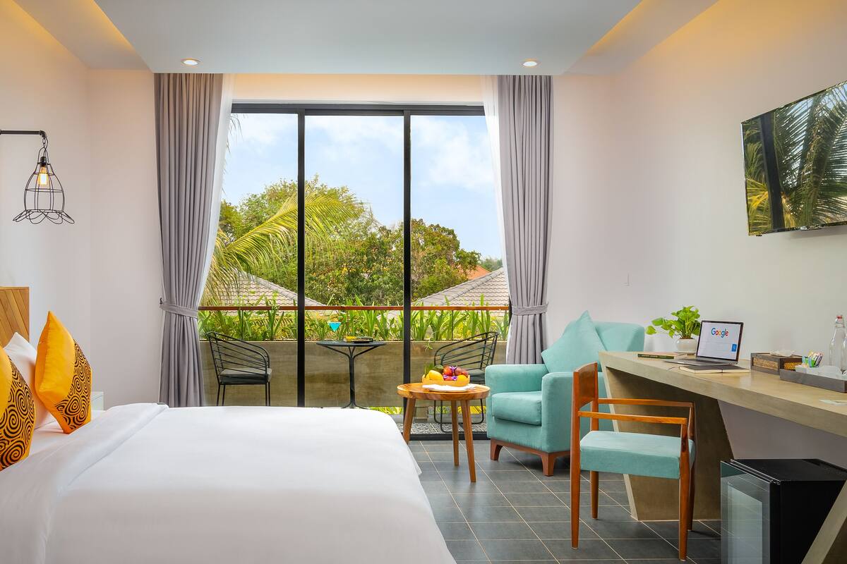 A hotel room with a bed, a desk with a laptop, a chair, a small table with fruit, and a balcony with a view of greenery.
