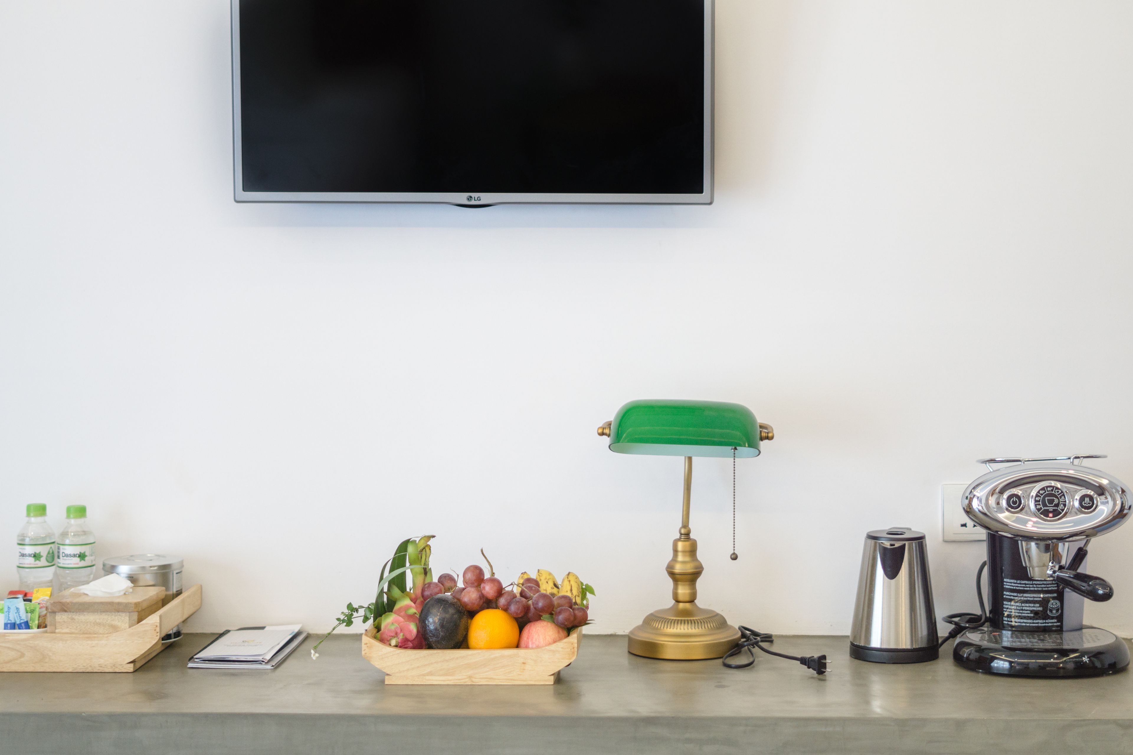 Une chambre moderne avec un téléviseur à écran plat fixé au mur, un panier à fruits en bois posé sur le comptoir, une lampe de bureau verte et une machine à café.