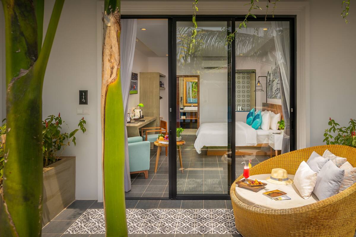A modern hotel room with a glass door leading to a balcony, a dining area, and a bedroom with a bed and pillows.