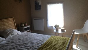 A bedroom with a bed, a chair, a small table, a radiator, and a window with curtains. - 2 Chambres Tramontane et Alizé (Jonquerettes)