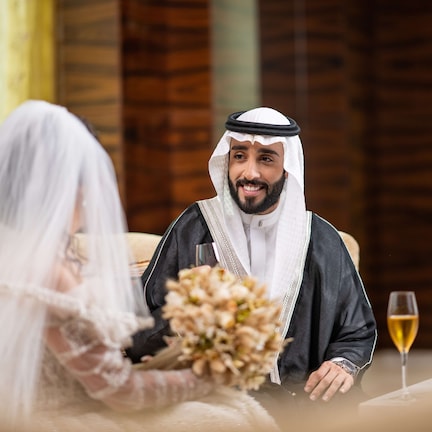 A man and a woman seated at a table with a glass of wine and a bouquet of flowers.