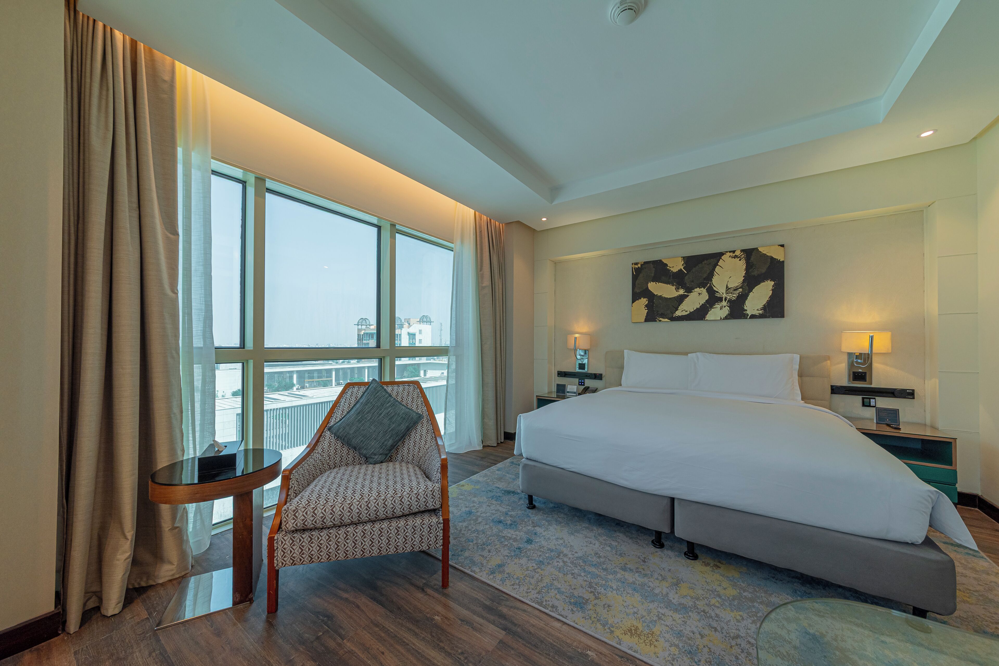 A hotel room with a large bed, a chair, a small table, a nightstand, and a window with curtains.