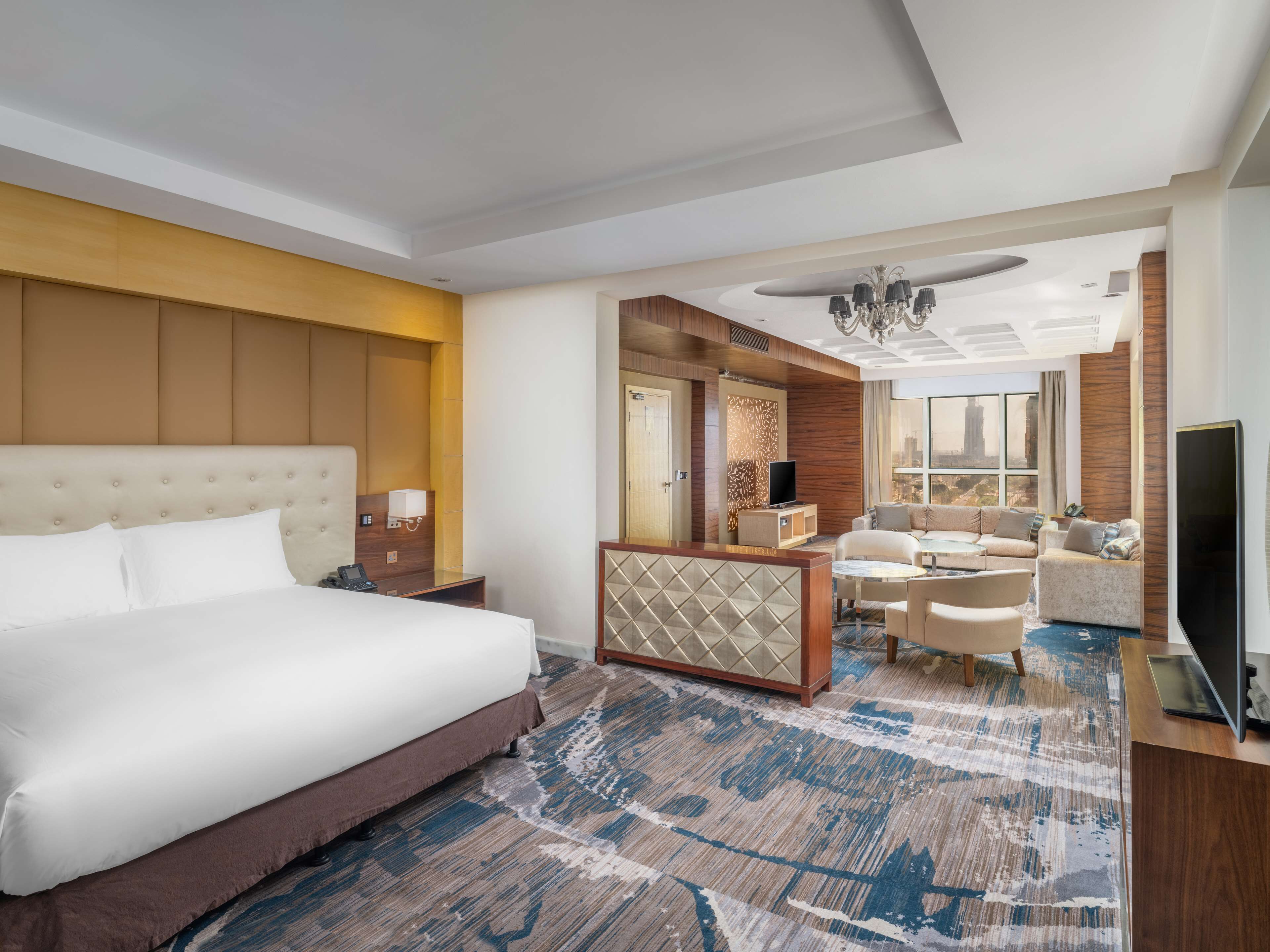 A hotel room with a large bed, a seating area with sofas, a TV, and a city view.