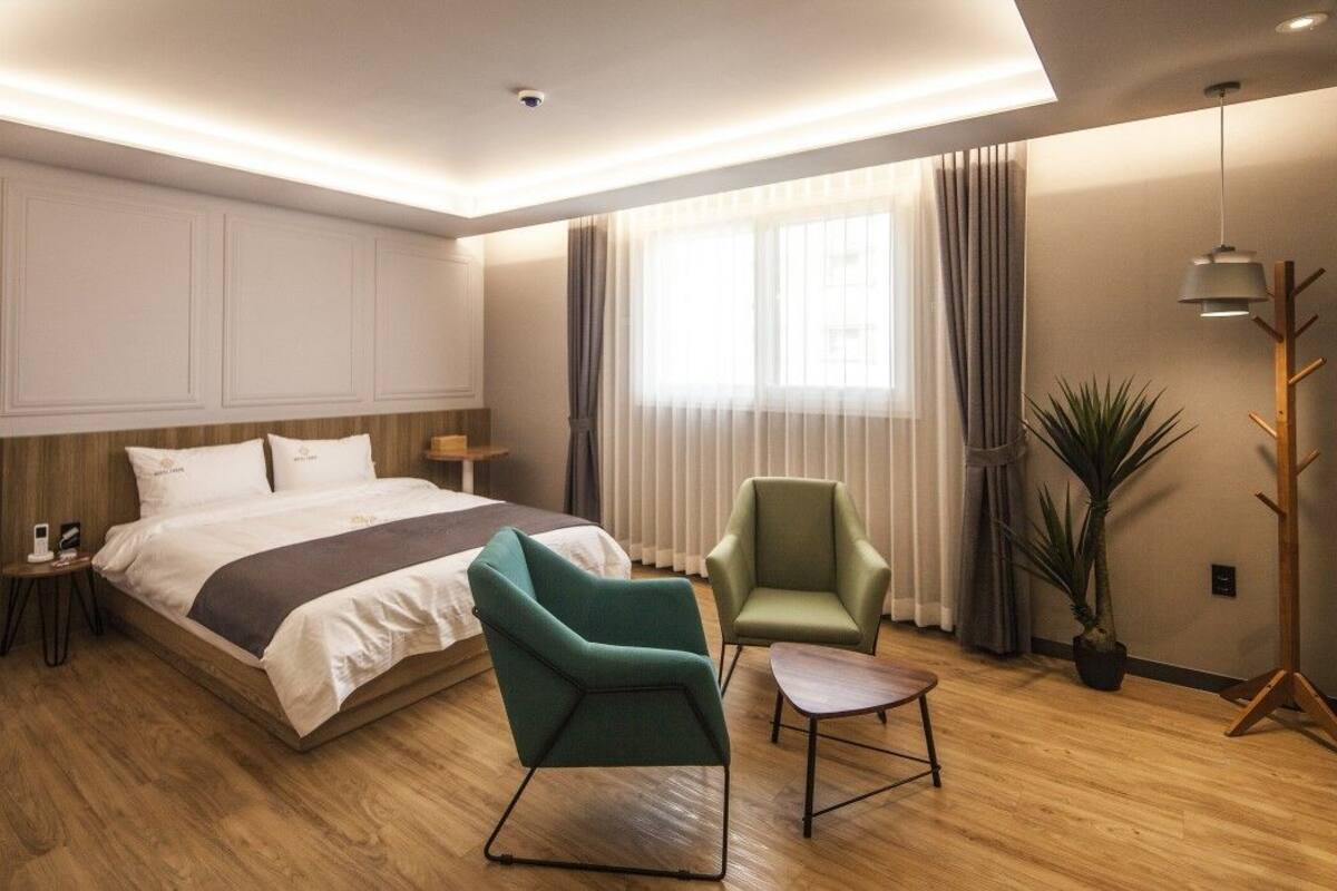 A modern hotel room with a large bed, two armchairs, a small table, and a potted plant.