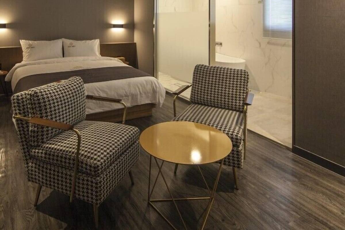 A hotel room with a bed, two checkered armchairs, a small round table, a sliding glass door, and a ceiling-mounted air vent.