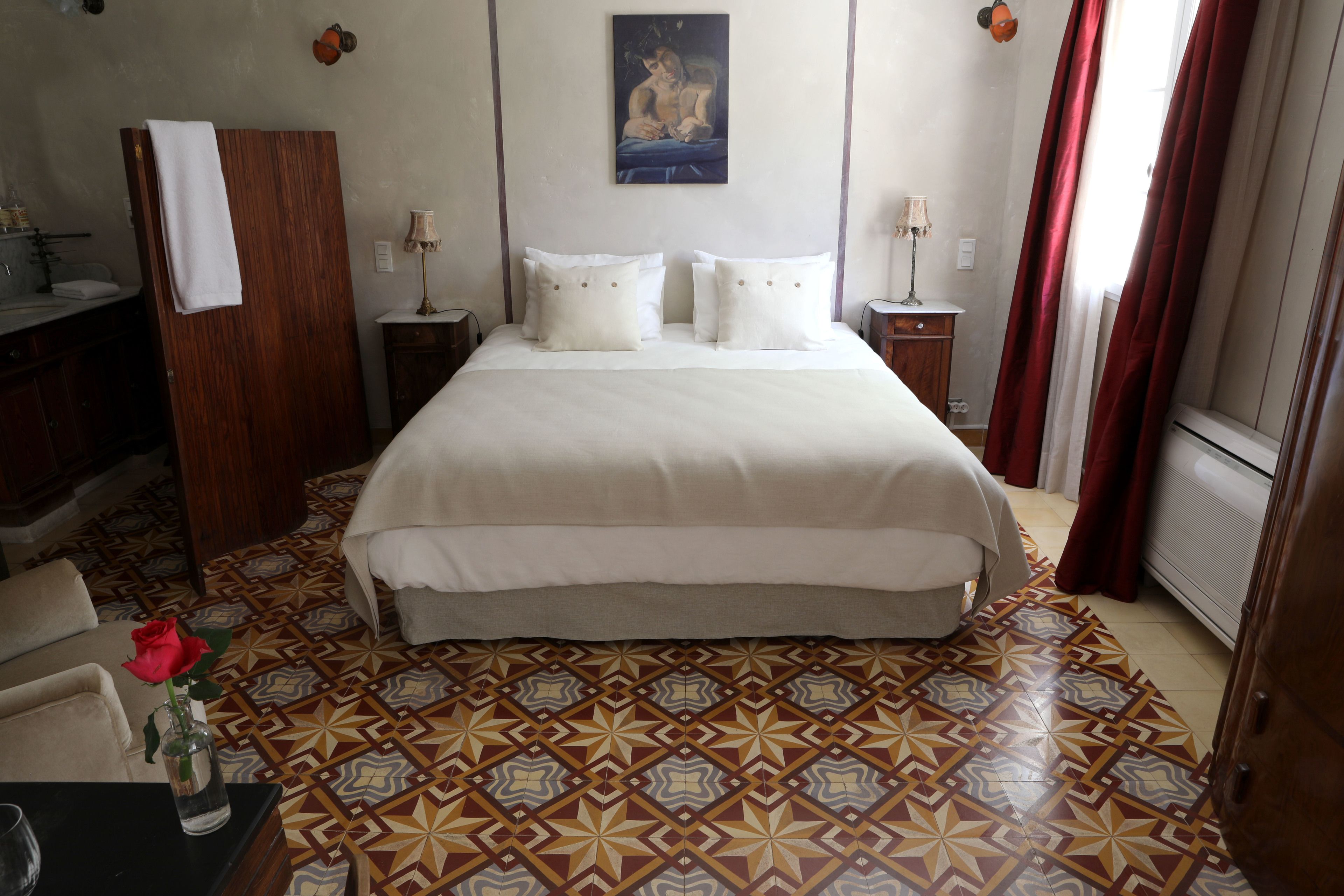 A hotel room with a large bed, a wooden wardrobe, a bathroom with a sink, and a patterned floor.