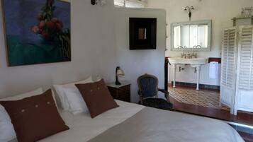 A bedroom with a large bed, a chair, a nightstand, a painting, a mirror, and a bathroom with a sink and mirror.