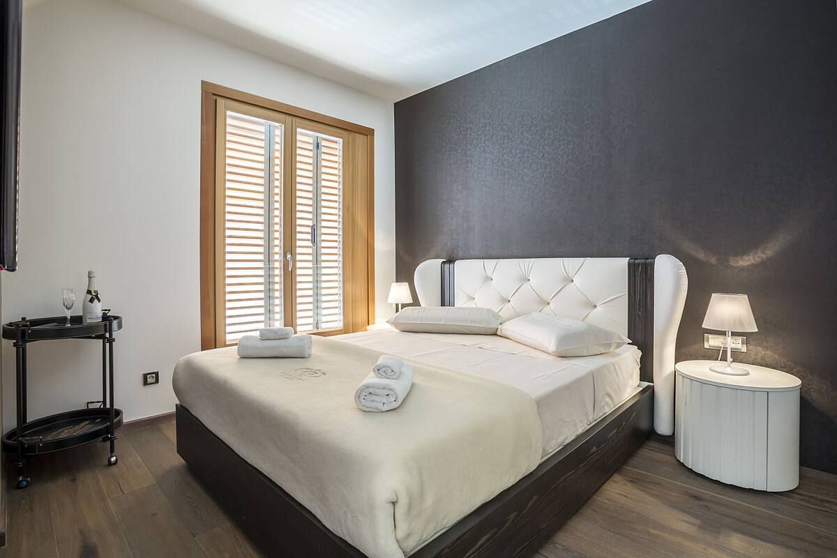 A bedroom with a large bed, bedside lamps, a nightstand, and a window with blinds.