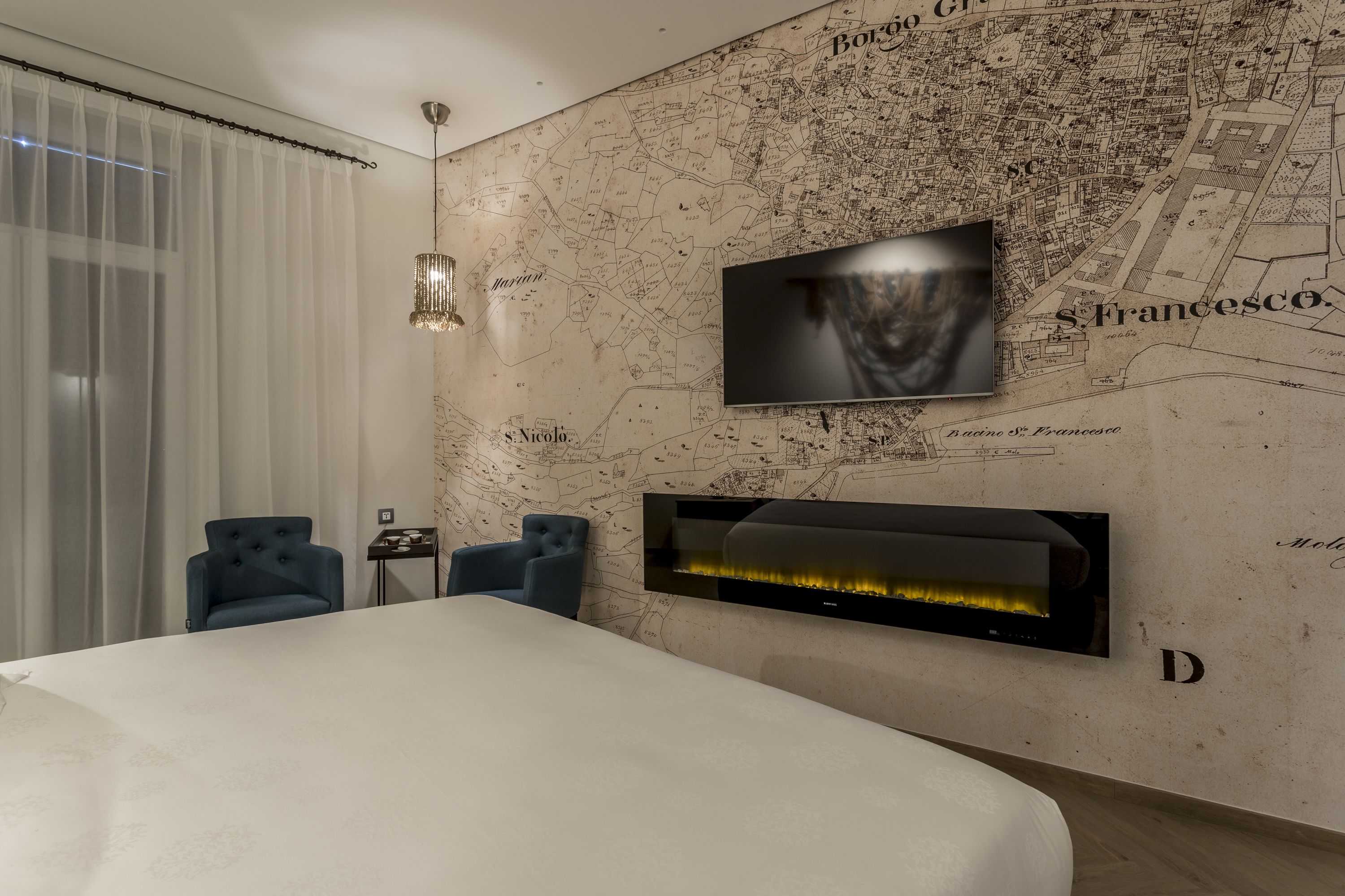 Une chambre moderne avec un grand lit, un téléviseur à écran plat fixé au mur, une cheminée et deux fauteuils bleus.