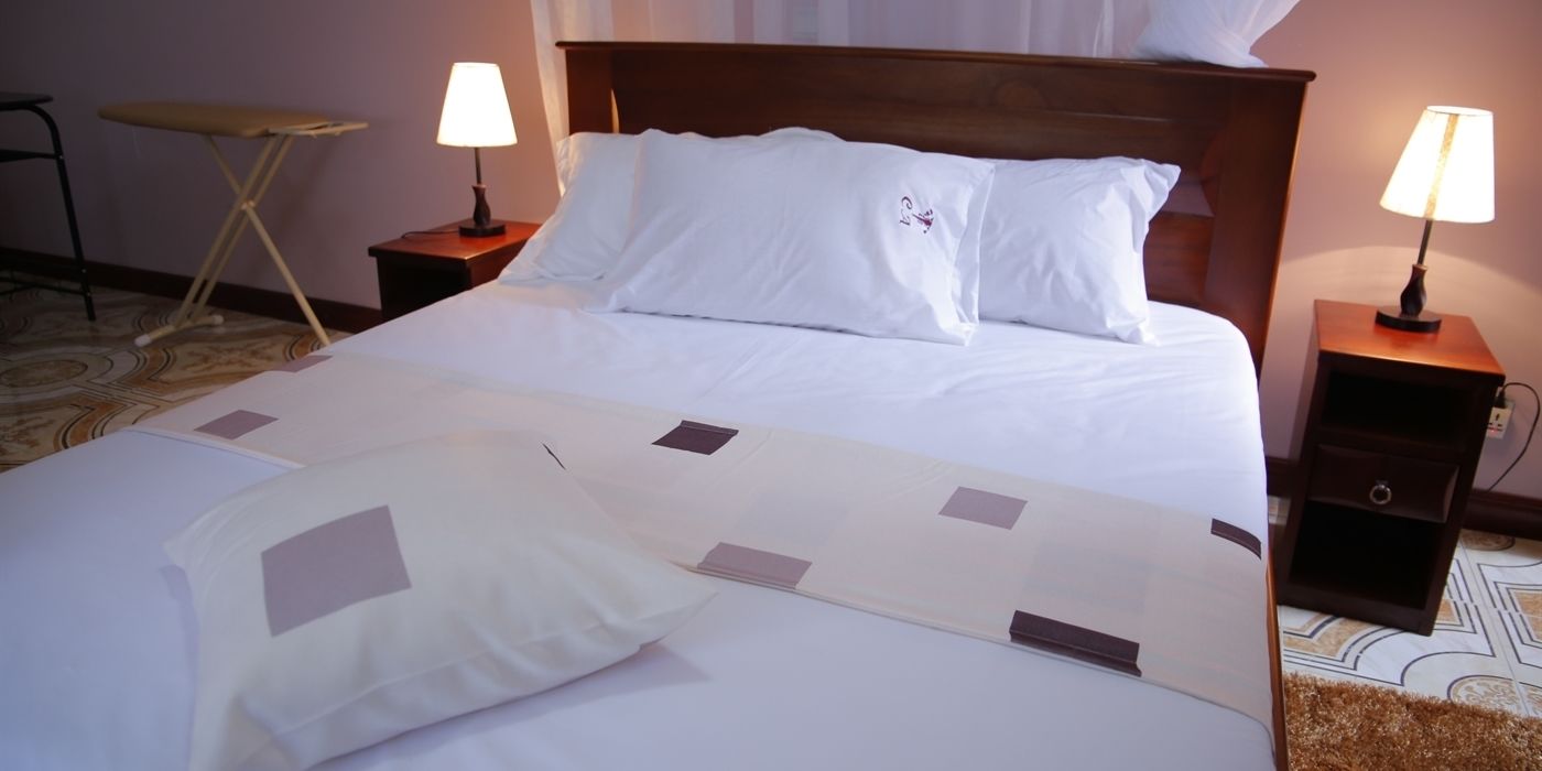 Uma cama bem arrumada com lençóis brancos e um edredom estampado, duas mesas de cabeceira com abajures e uma pequena mesa redonda no canto.