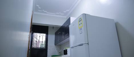 Cocina compacta con electrodomésticos blancos, incluyendo un refrigerador y un horno, y una ventana con celosías decorativas.