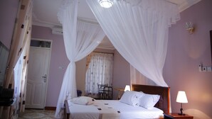 A bedroom with a canopy bed, a bedside table with a lamp, a chair, and a small table. - Crystal Hotel Apartments Entebbe (Entebbe)