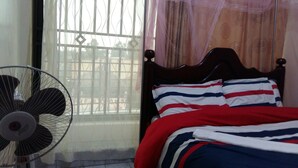 A room with a bed, a standing fan, and a window with a view of the outside.