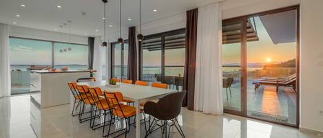 Un espace repas moderne avec une grande table et des chaises, de grandes fenêtres offrant une vue sur la mer, et un balcon d’où l’on peut admirer le coucher du soleil.