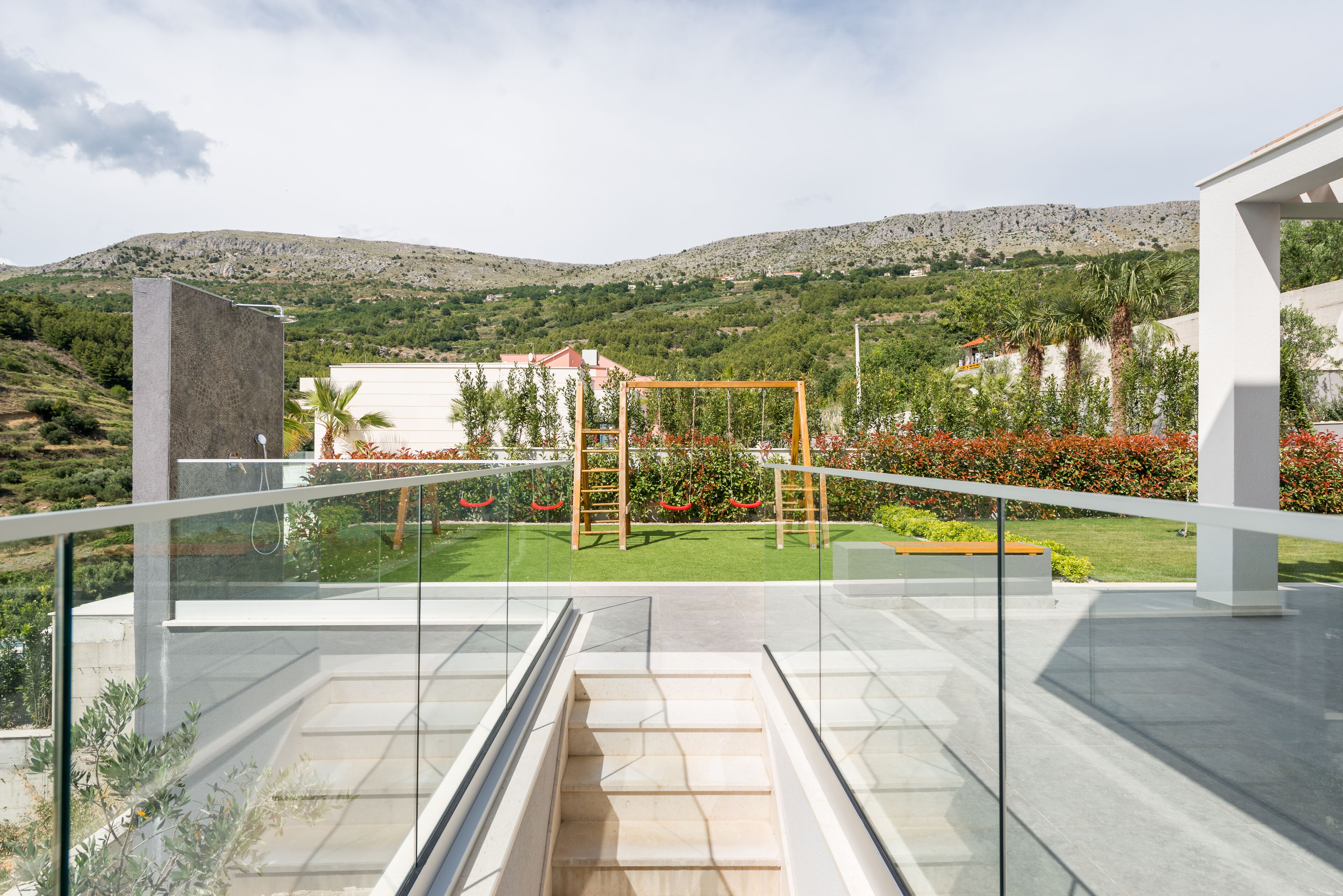 Une terrasse extérieure moderne, dotée d’une balustrade en verre, d’une balançoire en bois et offrant une vue sur un paysage montagneux.