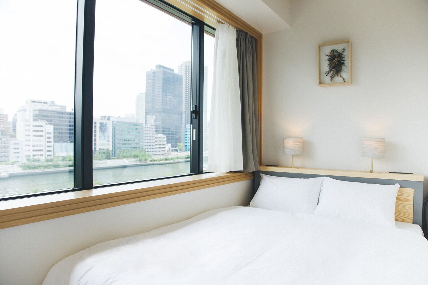 A hotel room with a large window, a bed, a nightstand with lamps, and a framed picture on the wall.