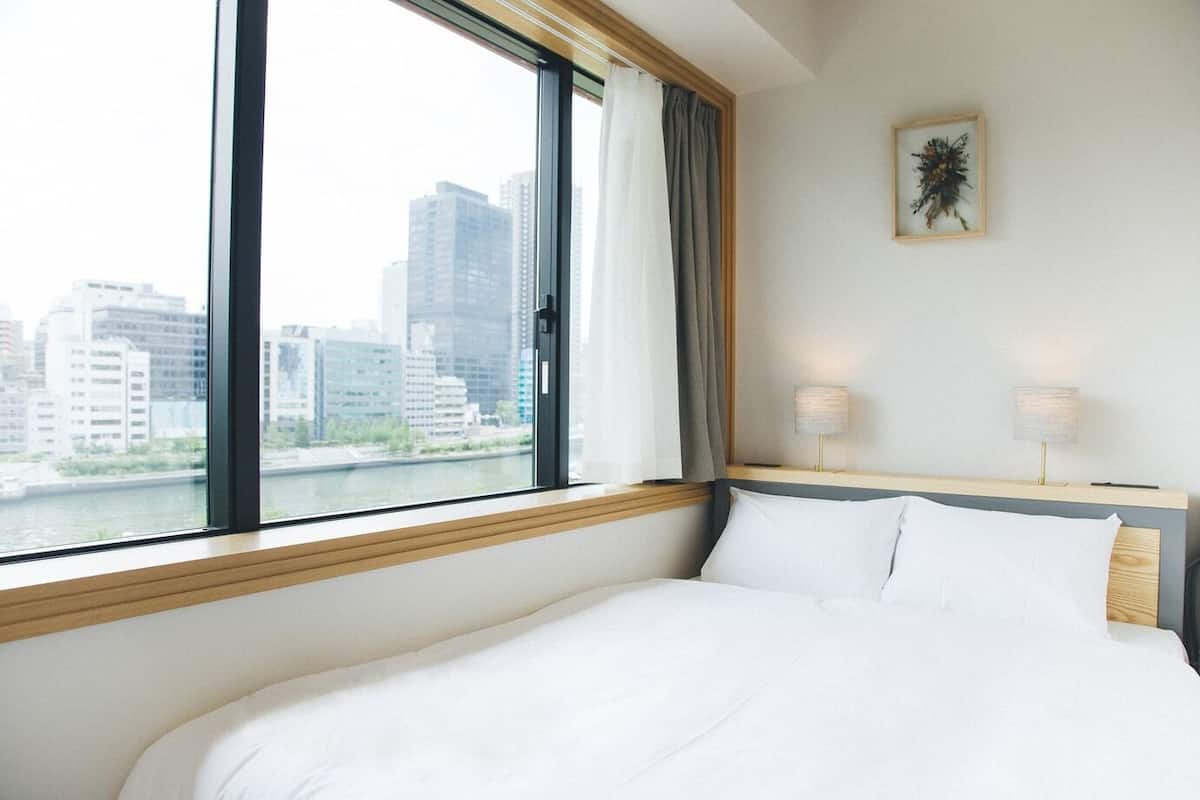 A hotel room with a large window, a bed, a nightstand with lamps, and a framed picture on the wall.