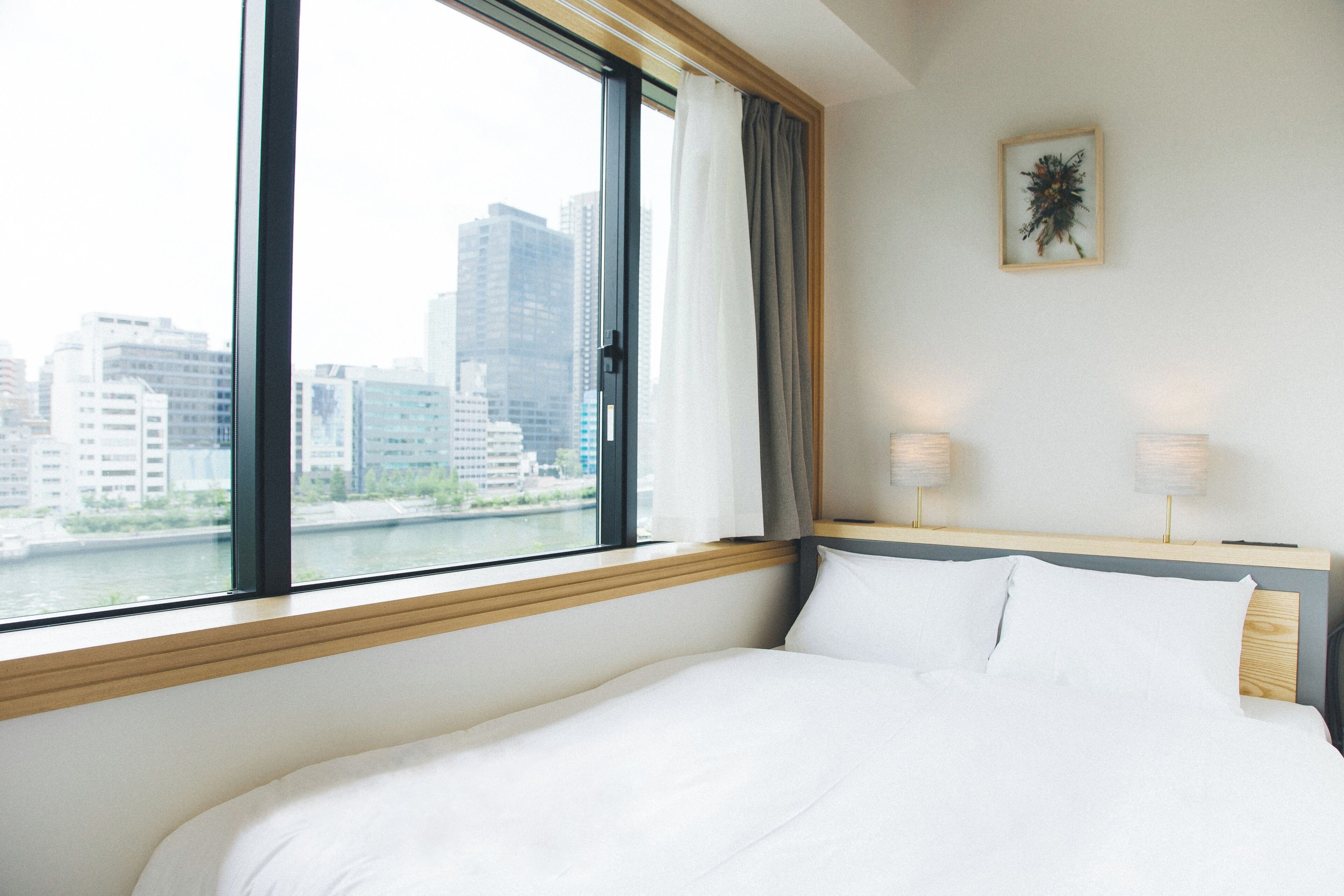 A hotel room with a large window, a bed, two bedside lamps, and a framed artwork on the wall.