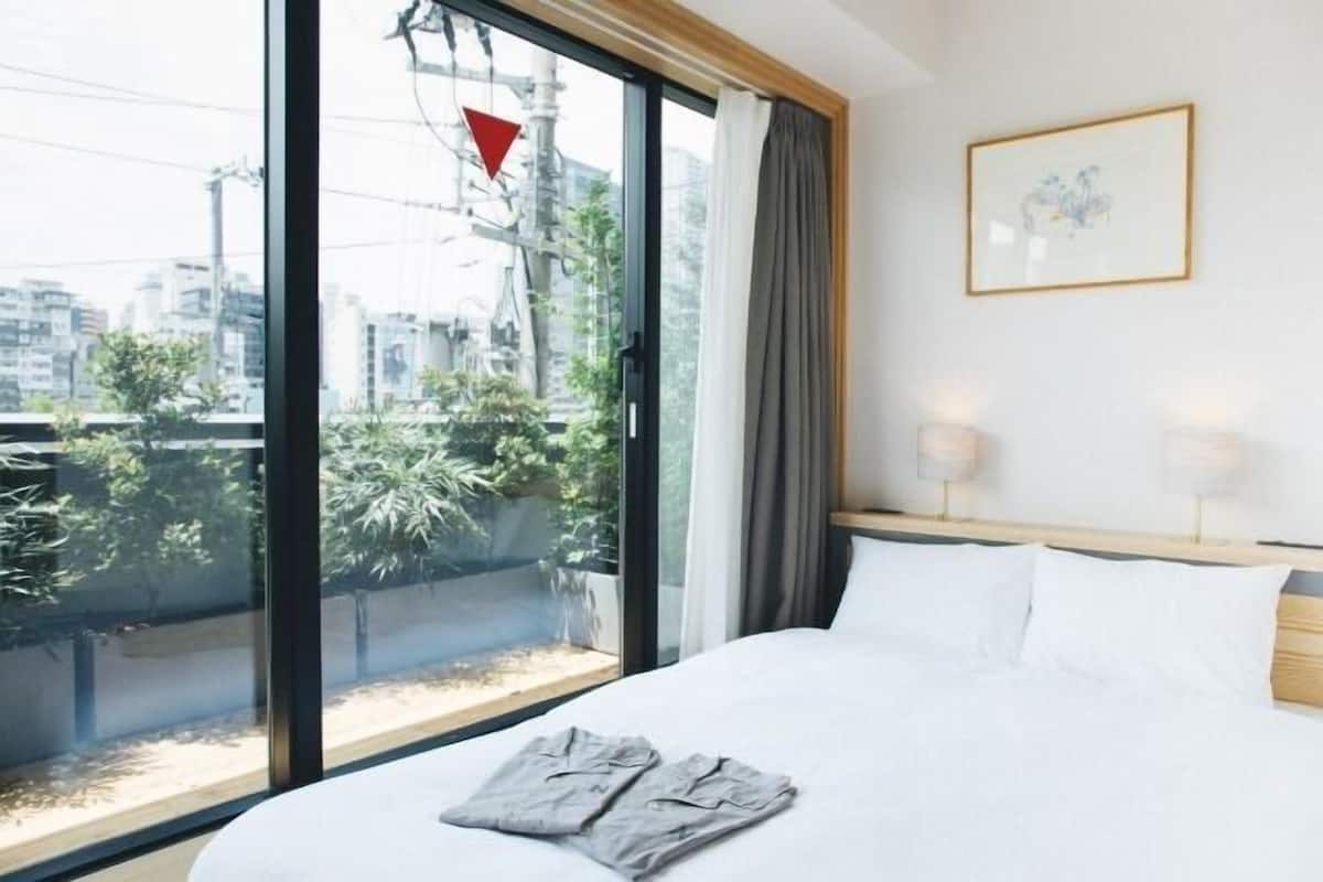 A hotel room with a large window, a bed with white linens, a framed picture on the wall, and a view of the cityscape.