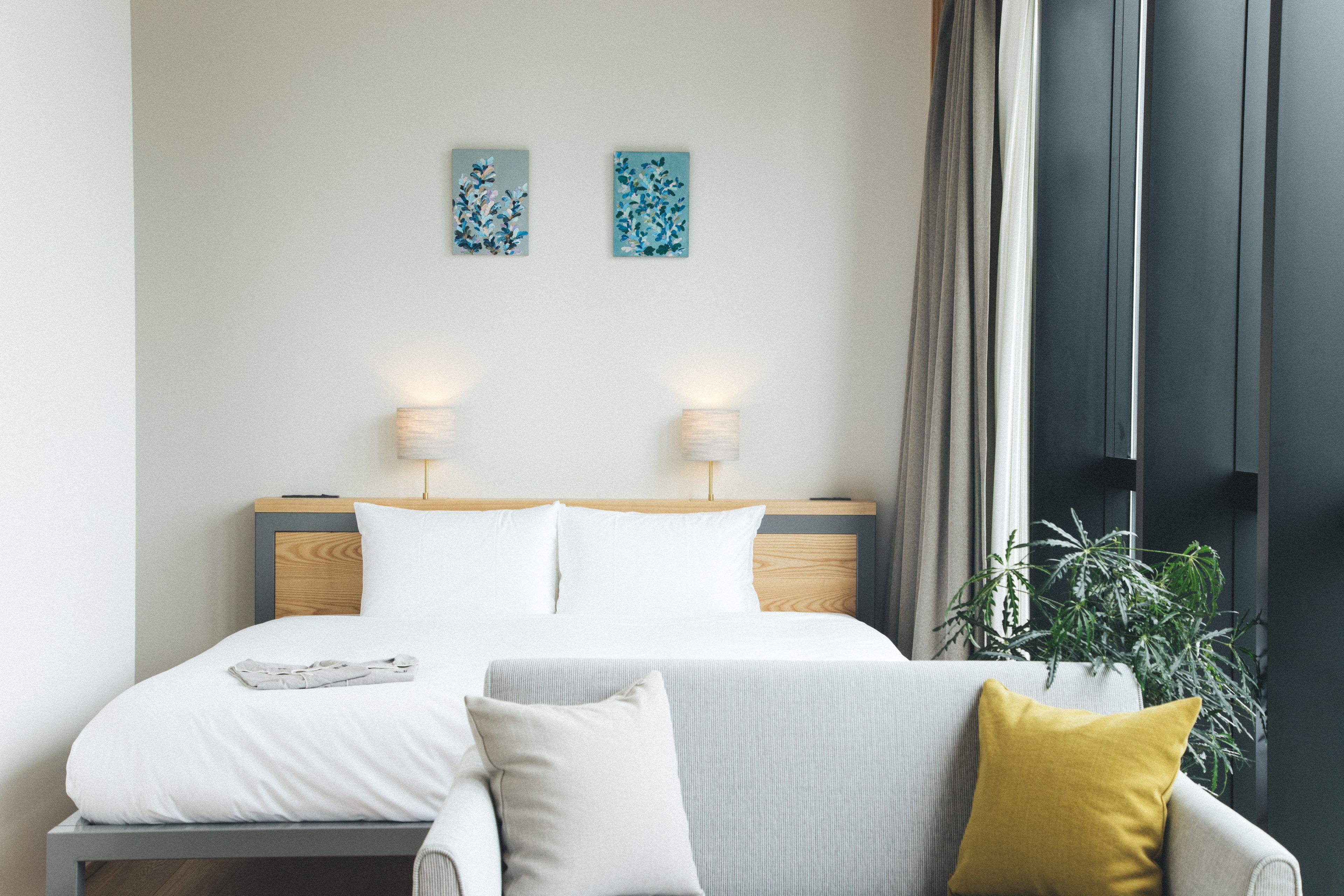 A modern hotel room with a neatly made bed, a grey sofa, and a potted plant.