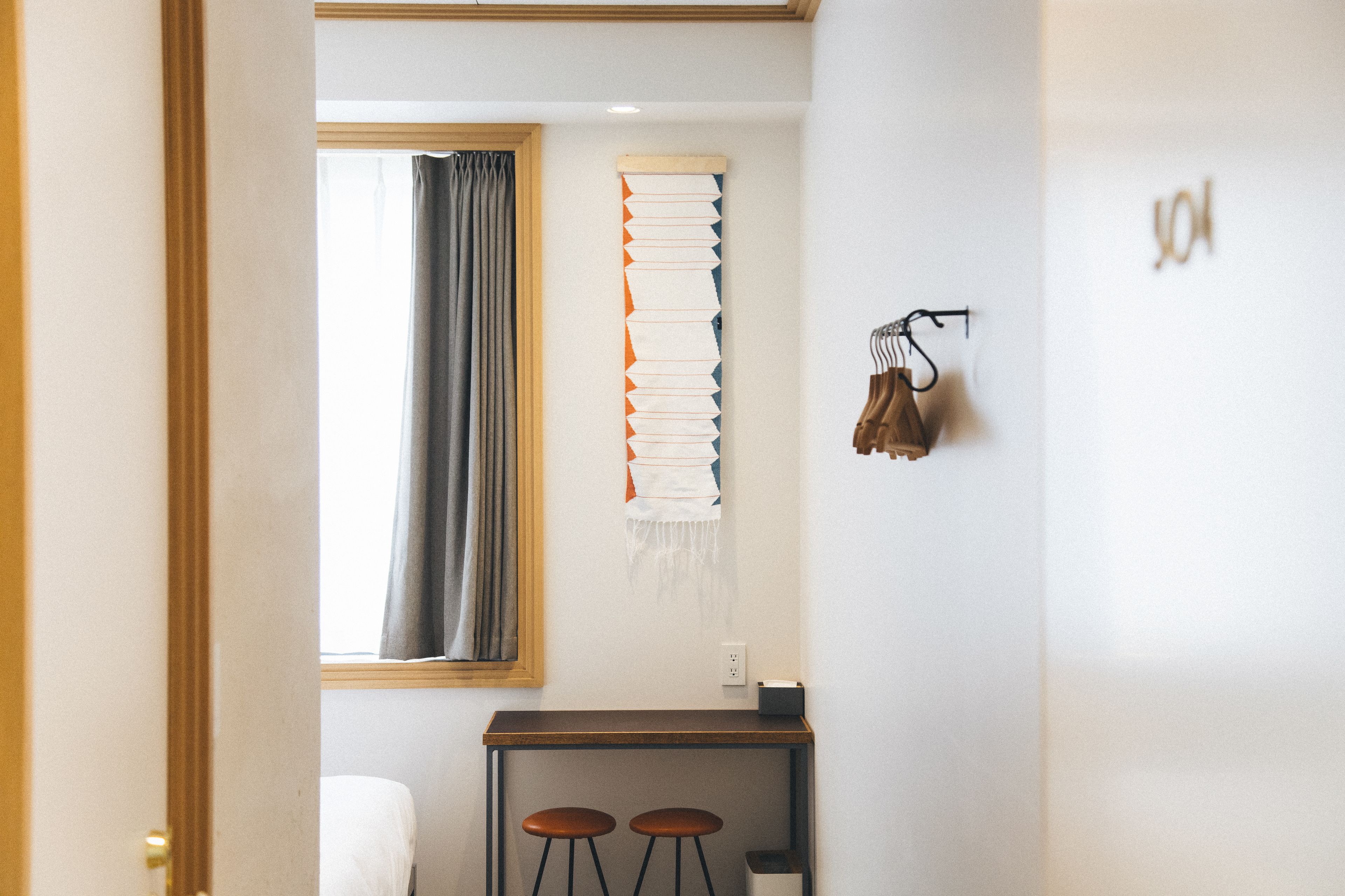 A hotel room with a bed, a small table with two stools, a window with curtains, and a wall-mounted shelf with books and a pair of glasses.