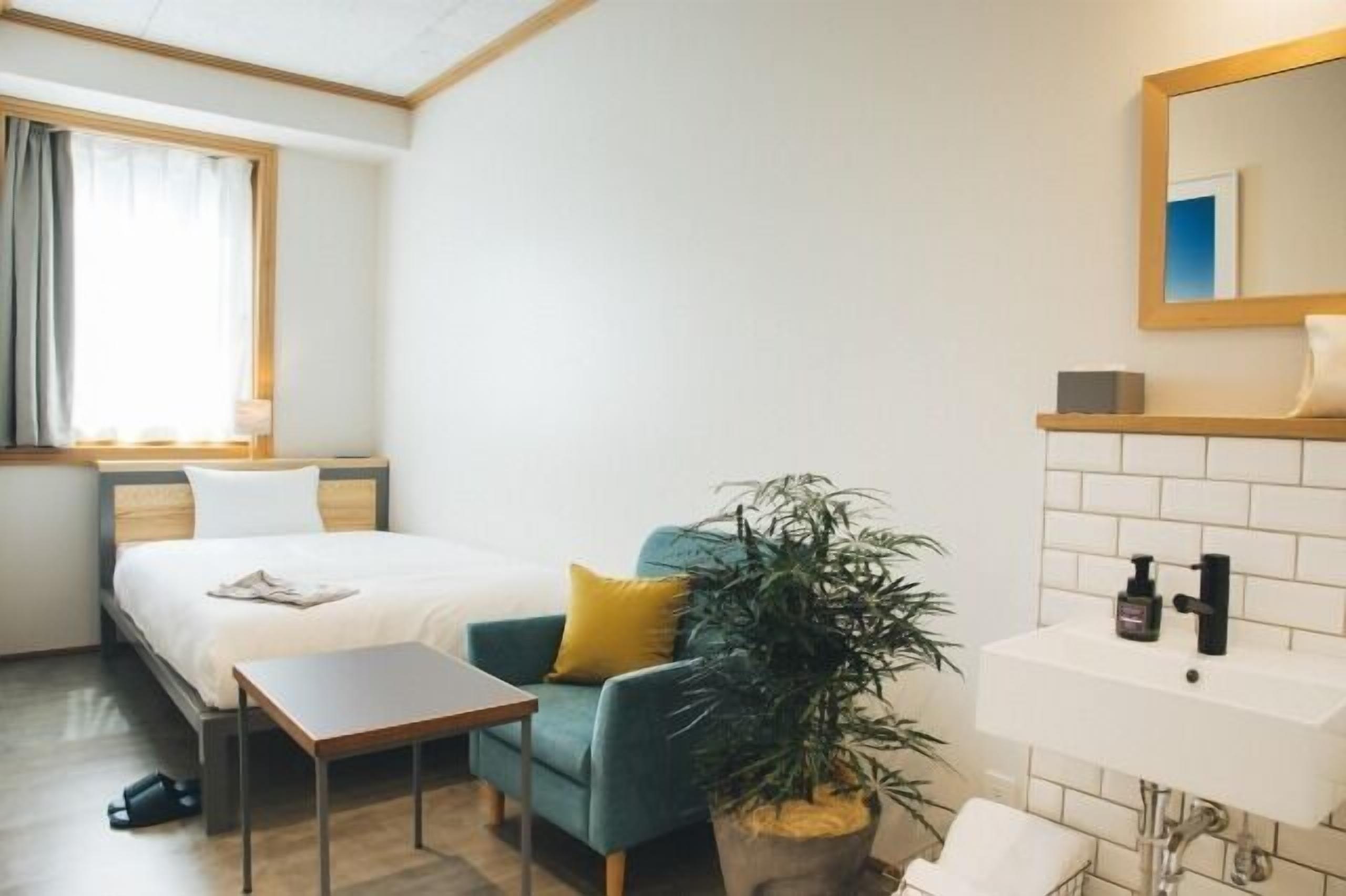 A hotel room with a bed, a small table, a chair, a potted plant, a sink, and a mirror.