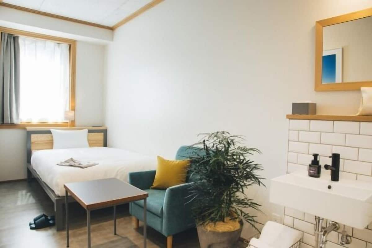A hotel room with a bed, a small table, a chair, a potted plant, a sink, and a mirror.