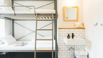 Una cama litera con escalera, un lavamanos pequeño y un espejo en una habitación con paredes blancas y suelo de baldosas.