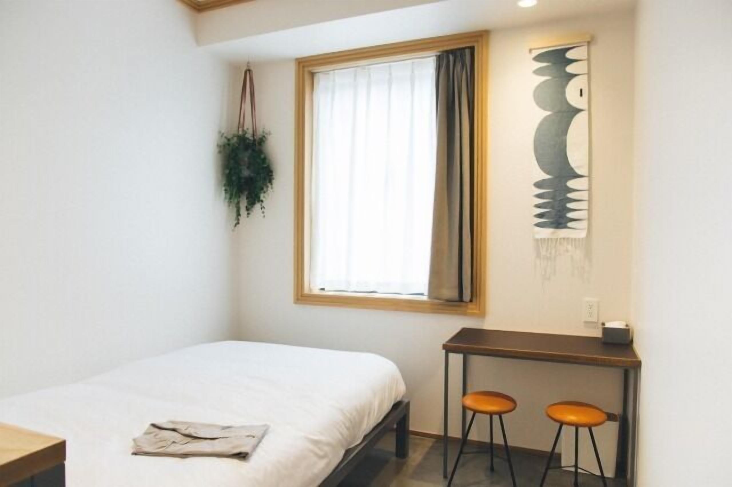 A small, clean room with a bed, a desk, two stools, a hanging plant, and a wall art piece.